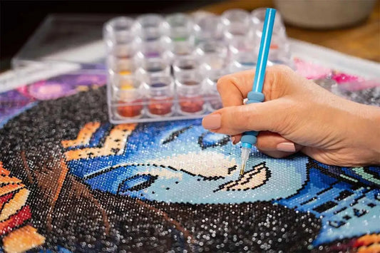 Hand die een pen vasthoudt en glinsterende zwarte steentjes aanbrengt op een diamond painting met blauwe en bruine tinten.