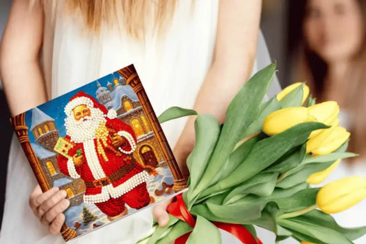 Vrouw in witte jurk houdt een boeket gele tulpen en een kerst diamond painting van de kerstman voor een besneeuwd gebouw.