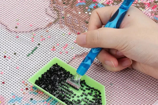 Hand met blauwe pen en groen bakje vol zwarte steentjes boven onvoltooide diamond painting met symbolen