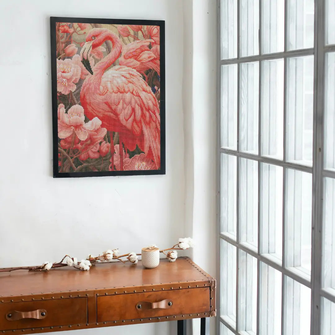 Opgehangen diamond painting van roze flamingo tussen bloemen aan witte muur bij groot raam en houten tafel
