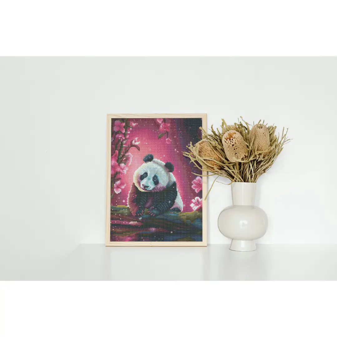 Ingelijste diamond painting van een panda met roze bloesems, geplaatst op een witte plank naast een vaas met gedroogde bloemen.