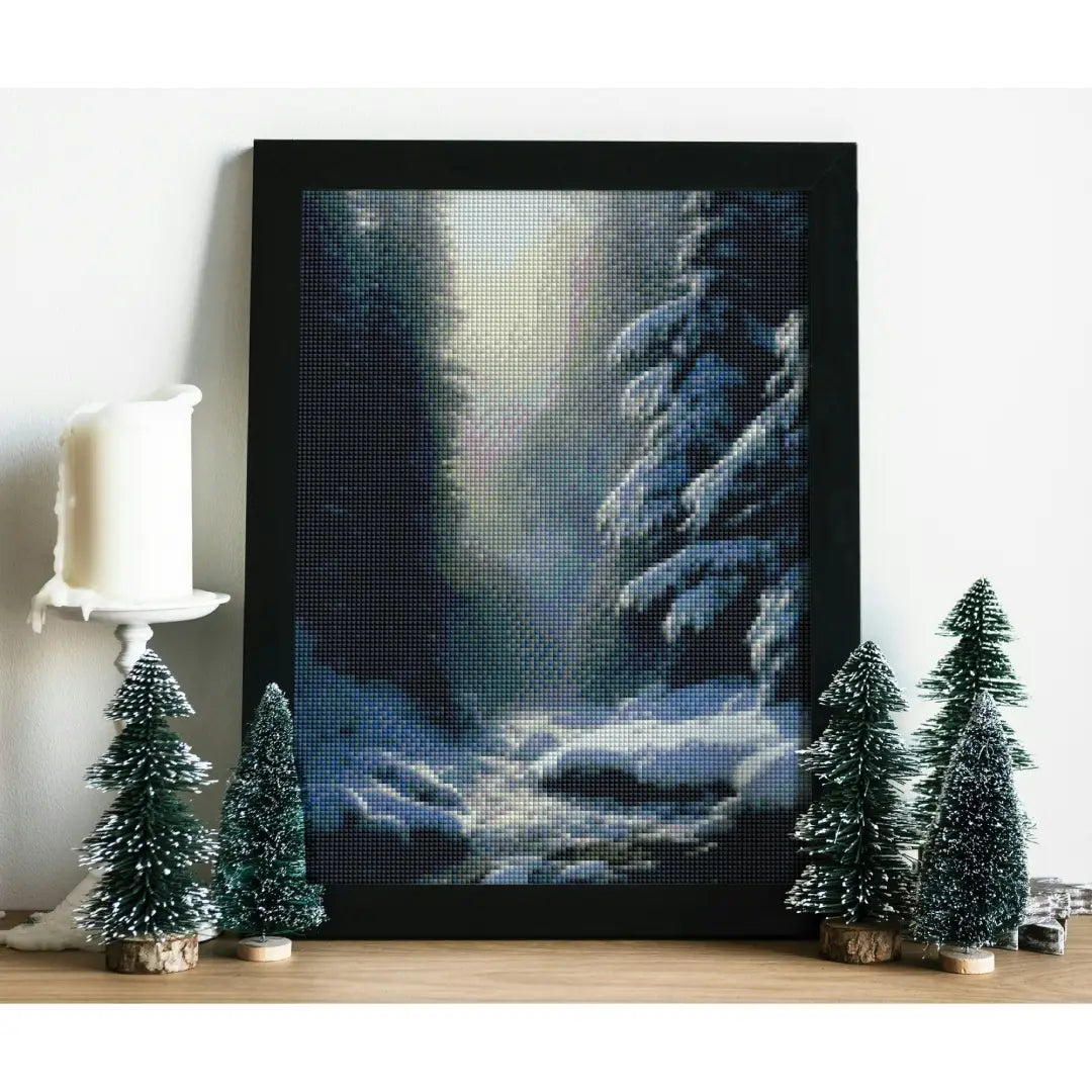 Diamond painting van een besneeuwd boslandschap, geplaatst op een oppervlak met kleine kerstboompjes en een witte kaars.