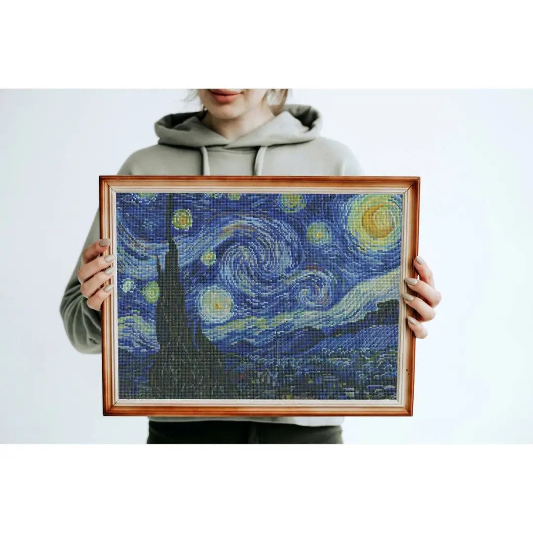 Ingelijste diamond painting geïnspireerd op De Sterrennacht met wervelende sterrenhemel, maan en dorp in blauwtinten, vastgehouden door een persoon.