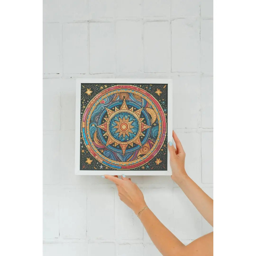 Ingelijste diamond painting van een mandala met een zon en sterren, vastgehouden door twee handen tegen een witte muur.