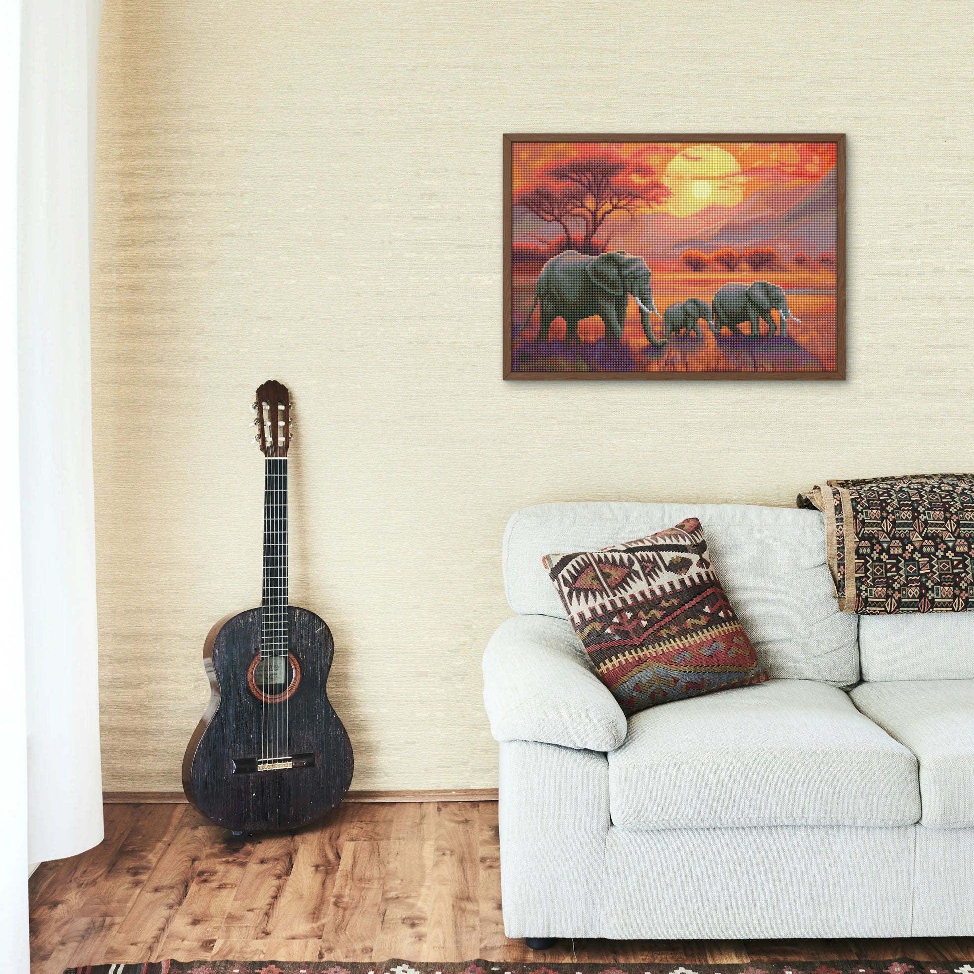 Diamond painting van een olifantenfamilie bij zonsondergang in een savannelandschap, ingelijst en opgehangen in een interieur met een gitaar en een lichte bank.